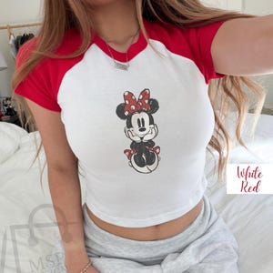 May include: White and red raglan crop top featuring a Minnie Mouse graphic. The shirt has a white body with red sleeves and neckline. The Minnie Mouse design is in black, red, and white. The text "White Red" is in a white box.