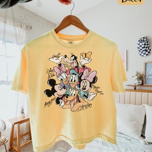 Puede incluir: Camiseta amarilla de corte cuadrado con un gr&aacute;fico de personajes de Disney, incluyendo Mickey Mouse, Minnie Mouse, Donald Duck, Daisy Duck, Goofy y Pluto. La camiseta tiene el texto "COMFORT COLORS - BOXY TEE" y "Butter".
