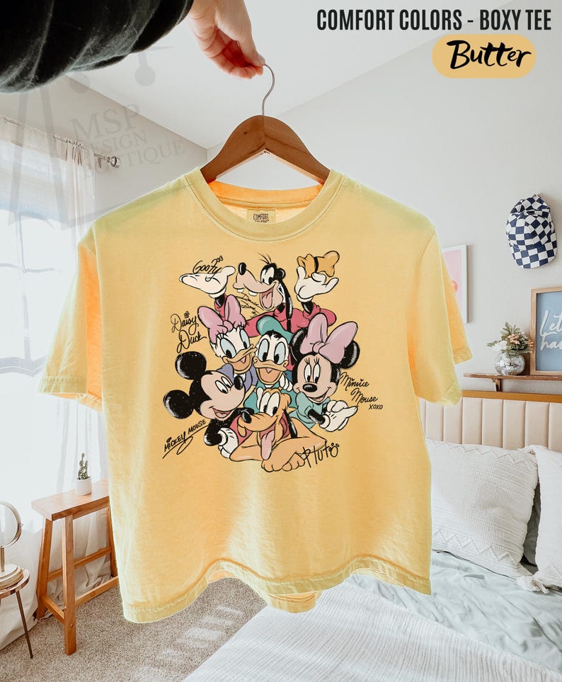 Puede incluir: Camiseta amarilla de corte cuadrado con un gr&aacute;fico de personajes de Disney, incluyendo Mickey Mouse, Minnie Mouse, Donald Duck, Daisy Duck, Goofy y Pluto. La camiseta tiene el texto "COMFORT COLORS - BOXY TEE" y "Butter".