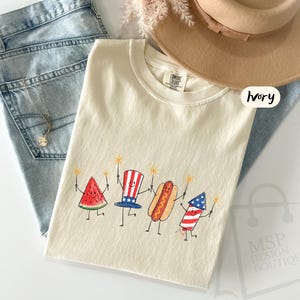 May include: An ivory short-sleeve t-shirt featuring a graphic print of a smiling watermelon slice, a top hat with stars and stripes, a hot dog, and a firework, all holding sparklers. The shirt has a Comfort Colors label and the text Ivory.