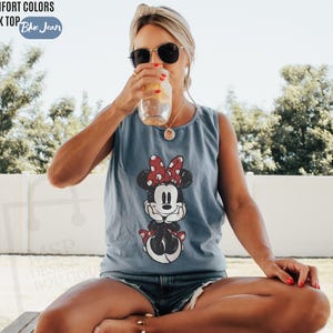 May include: A blue jean colored tank top featuring a graphic print of Minnie Mouse with a red and white polka dot bow. The text Comfort Colors Tank Top Blue Jean is visible in the upper left corner of the image, along with a faint watermark.