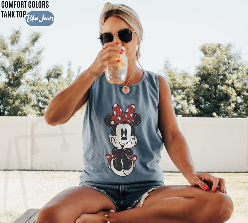 May include: A blue jean colored tank top featuring a graphic print of Minnie Mouse with a red and white polka dot bow. The text Comfort Colors Tank Top Blue Jean is visible in the upper left corner of the image, along with a faint watermark.