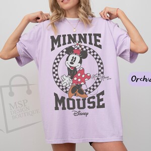 May include: Orchid-colored t-shirt featuring a Minnie Mouse graphic. The design includes the words "MINNIE MOUSE" in bold, black letters, with Minnie Mouse in the center. The Disney logo is at the bottom.