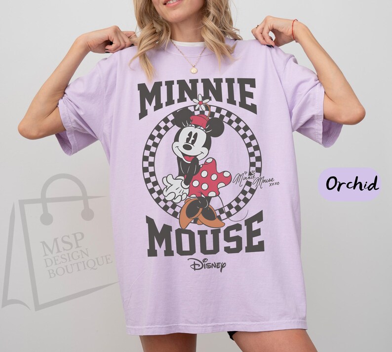 May include: Orchid-colored t-shirt featuring a Minnie Mouse graphic. The design includes the words "MINNIE MOUSE" in bold, black letters, with Minnie Mouse in the center. The Disney logo is at the bottom.