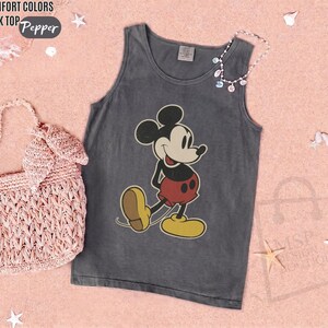 May include: Gray tank top featuring a classic Mickey Mouse graphic. The sleeveless shirt has a vintage look, with a distressed appearance. The image also includes a pink woven handbag and a necklace with colorful beads. The words "COMFORT COLORS TANK TOP Pepper" are in the top left corner.