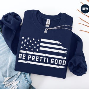 May include: Navy blue sweatshirt with a distressed American flag design. The flag features white stars and stripes, with the text "BE PRETTI GOOD" in white. A silver necklace with a pendant is draped across the sweatshirt.