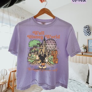 May include: Orchid-colored boxy tee with a graphic of Walt Disney World, featuring Mickey Mouse, a castle, and Spaceship Earth. The text "Walt Disney World" is in orange. The shirt is hanging on a wooden hanger.