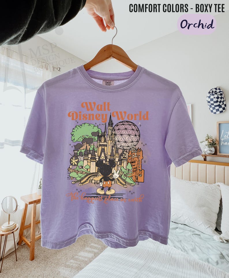 May include: Orchid-colored boxy tee with a graphic of Walt Disney World, featuring Mickey Mouse, a castle, and Spaceship Earth. The text "Walt Disney World" is in orange. The shirt is hanging on a wooden hanger.