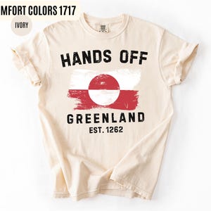 May include: Ivory-colored t-shirt with the text "HANDS OFF GREENLAND" in black, along with a Greenlandic flag design. The shirt also features "EST. 1262" in black. The shirt is made of a soft material.
