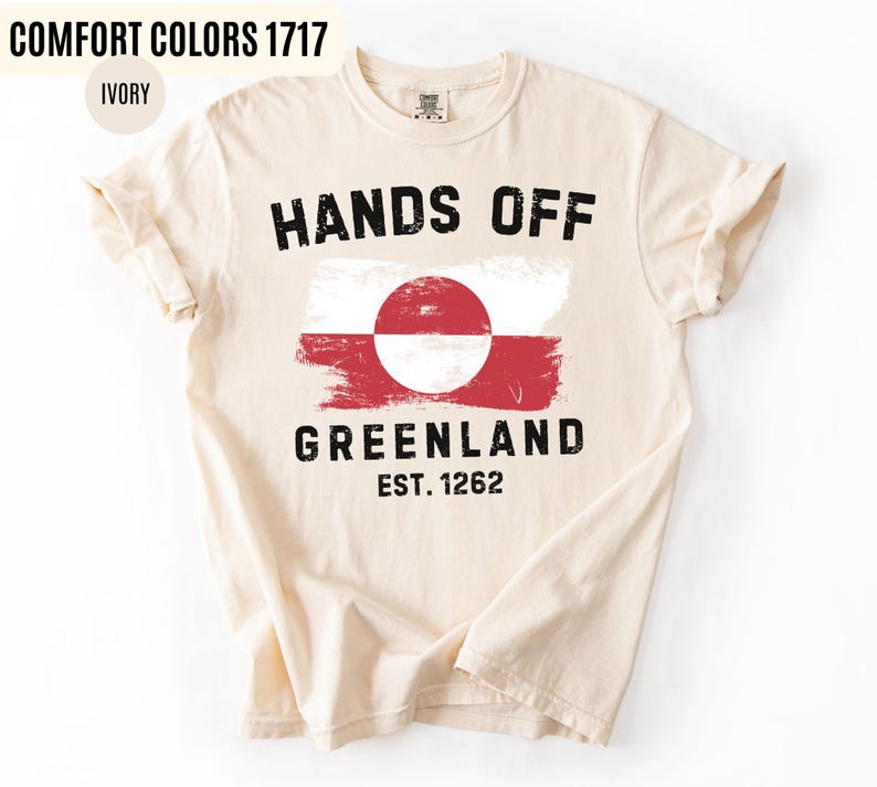 May include: Ivory-colored t-shirt with the text "HANDS OFF GREENLAND" in black, along with a Greenlandic flag design. The shirt also features "EST. 1262" in black. The shirt is made of a soft material.