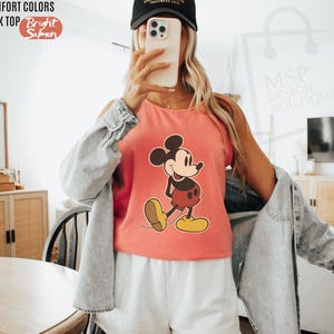 May include: A bright salmon-colored tank top featuring a classic Mickey Mouse graphic. The tank top has the words "COMFORT COLORS" and "TANK TOP" printed at the top left. The words "Bright Salmon" are printed in a circle at the top left.