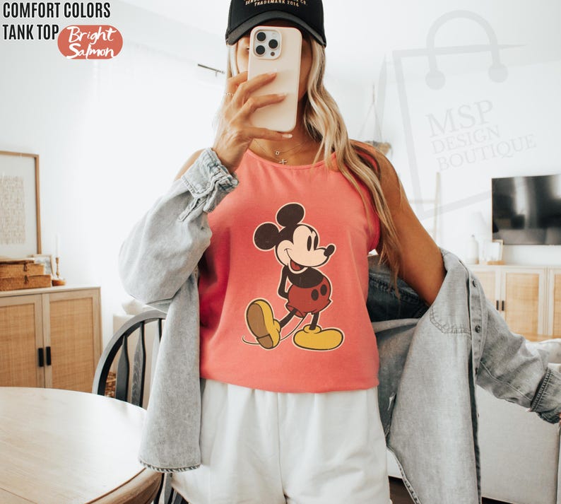 May include: A bright salmon-colored tank top featuring a classic Mickey Mouse graphic. The tank top has the words "COMFORT COLORS" and "TANK TOP" printed at the top left. The words "Bright Salmon" are printed in a circle at the top left.