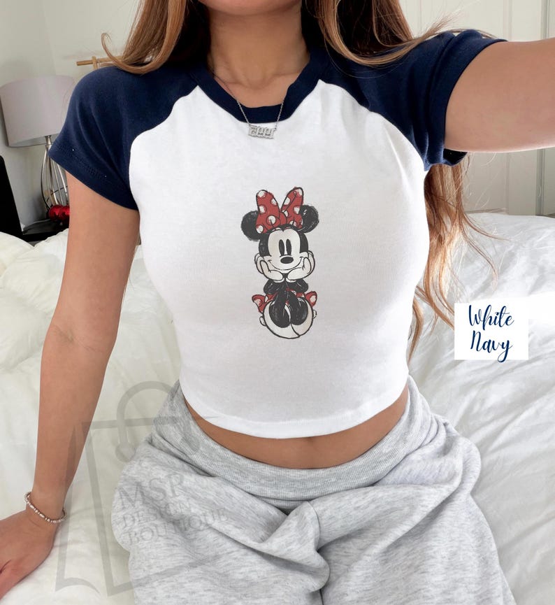 May include: White crop top with navy blue short sleeves and a Minnie Mouse graphic. The graphic features Minnie Mouse with a red bow and polka dots. The text "White Navy" is visible on the right side.