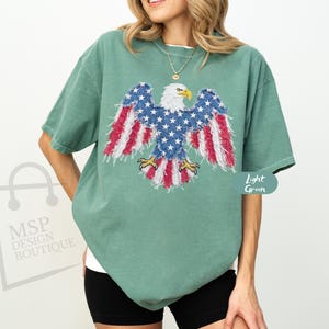 May include: A light green short-sleeve t-shirt featuring a graphic of a bald eagle with wings patterned in red, white, and blue stars and stripes. The eagle has a white head, yellow beak, and yellow talons, displayed centrally on the chest.