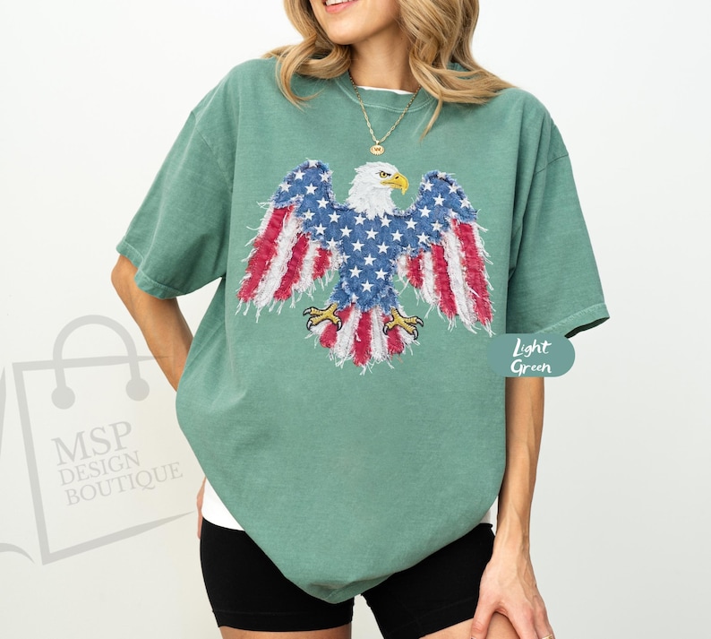 May include: A light green short-sleeve t-shirt featuring a graphic of a bald eagle with wings patterned in red, white, and blue stars and stripes. The eagle has a white head, yellow beak, and yellow talons, displayed centrally on the chest.