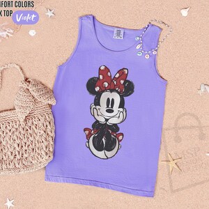 May include: A violet tank top featuring a graphic print of Minnie Mouse with a red and white polka dot bow. The tank top has a crew neckline and a Comfort Colors brand label at the inner collar. A beige crocheted handbag is positioned to the left.