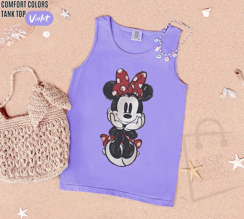 May include: A violet tank top featuring a graphic print of Minnie Mouse with a red and white polka dot bow. The tank top has a crew neckline and a Comfort Colors brand label at the inner collar. A beige crocheted handbag is positioned to the left.