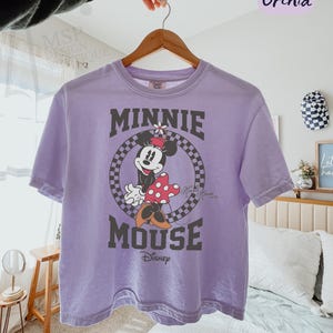 以下が含まれることがあります： ミニーマウスのグラフィックが特徴のラベンダー色のComfort ColorsボックスTシャツ。「MINNIE MOUSE」の文字が円形の市松模様でデザインされ、下にディズニーのロゴが入っています。