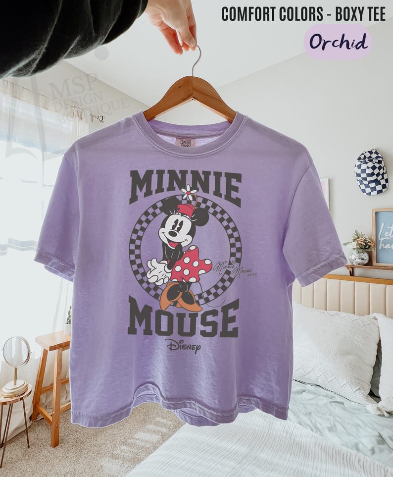 以下が含まれることがあります： ミニーマウスのグラフィックが特徴のラベンダー色のComfort ColorsボックスTシャツ。「MINNIE MOUSE」の文字が円形の市松模様でデザインされ、下にディズニーのロゴが入っています。