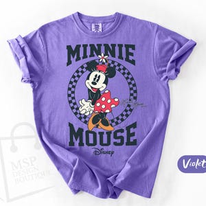 May include: Violet-colored t-shirt featuring a graphic of Minnie Mouse. The design includes the text "MINNIE MOUSE" in black block letters, with Minnie Mouse in the center. She is wearing a red and white polka dot dress and a flower on her head. The Disney logo is at the bottom.