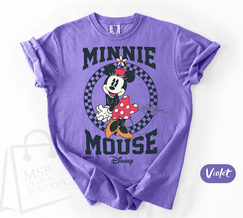 May include: Violet-colored t-shirt featuring a graphic of Minnie Mouse. The design includes the text "MINNIE MOUSE" in black block letters, with Minnie Mouse in the center. She is wearing a red and white polka dot dress and a flower on her head. The Disney logo is at the bottom.