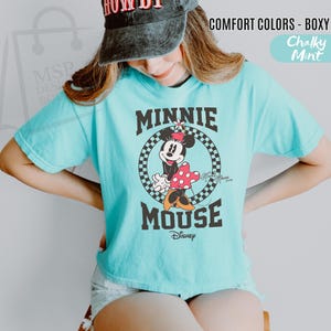 以下が含まれることがあります： 水色のTシャツ。チェック柄の円の中にミニーマウスのグラフィックがあり、その上下に「MINNIE MOUSE」の文字がプリントされています。ディズニーのロゴも表示されています。