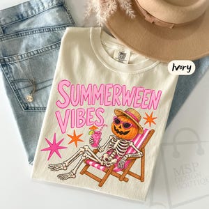 May include: An ivory t-shirt featuring a graphic of a skeleton with a pumpkin head wearing sunglasses and a straw hat, lounging in a pink and white striped beach chair while holding a pink drink. The text reads Summerween Vibes in pink.