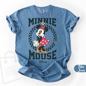 May include: Blue jean t-shirt featuring a graphic of Minnie Mouse with the text "MINNIE MOUSE" in a collegiate style font. Minnie is wearing a red and white polka dot dress and a flower in her hair. The Disney logo is visible.