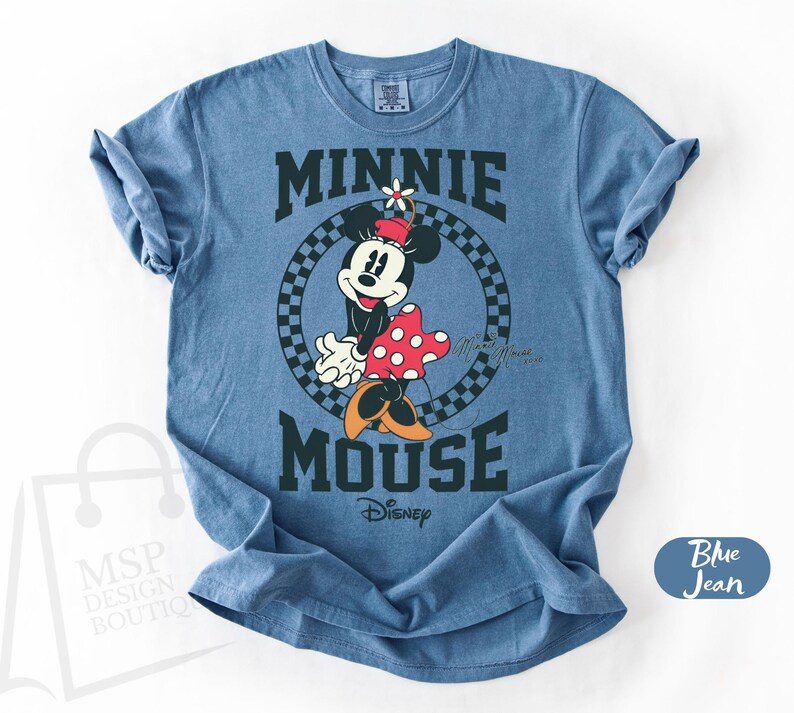 May include: Blue jean t-shirt featuring a graphic of Minnie Mouse with the text "MINNIE MOUSE" in a collegiate style font. Minnie is wearing a red and white polka dot dress and a flower in her hair. The Disney logo is visible.