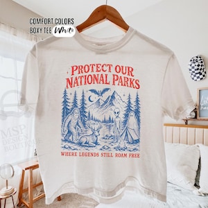 May include: A white boxy t-shirt featuring a graphic print in red and blue. The design includes mountains, pine trees, a bat, a deer, a ghost, an alien, and Bigfoot, with the text "PROTECT OUR NATIONAL PARKS" and "WHERE LEGENDS STILL ROAM FREE."