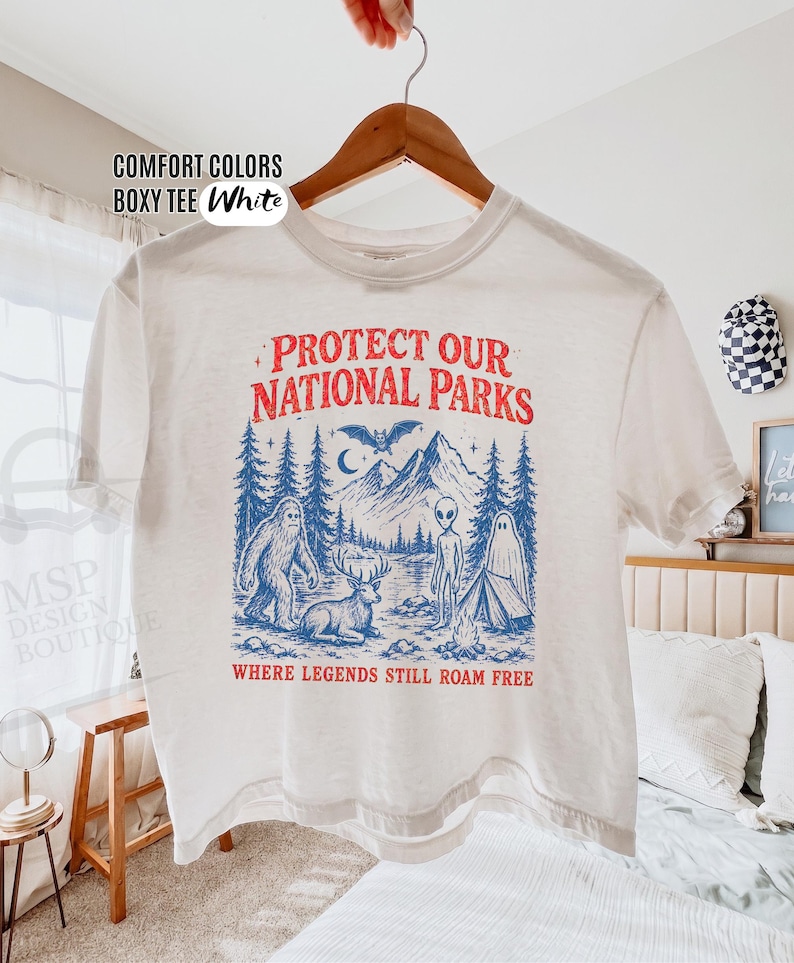 May include: A white boxy t-shirt featuring a graphic print in red and blue. The design includes mountains, pine trees, a bat, a deer, a ghost, an alien, and Bigfoot, with the text "PROTECT OUR NATIONAL PARKS" and "WHERE LEGENDS STILL ROAM FREE."