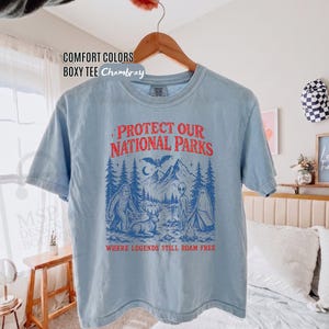 May include: A light blue short-sleeve t-shirt with a graphic print in red and blue. The text reads "PROTECT OUR NATIONAL PARKS" and "WHERE LEGENDS STILL ROAM FREE" above and below an illustration of mountains, a forest, a deer, a ghost, and an alien.
