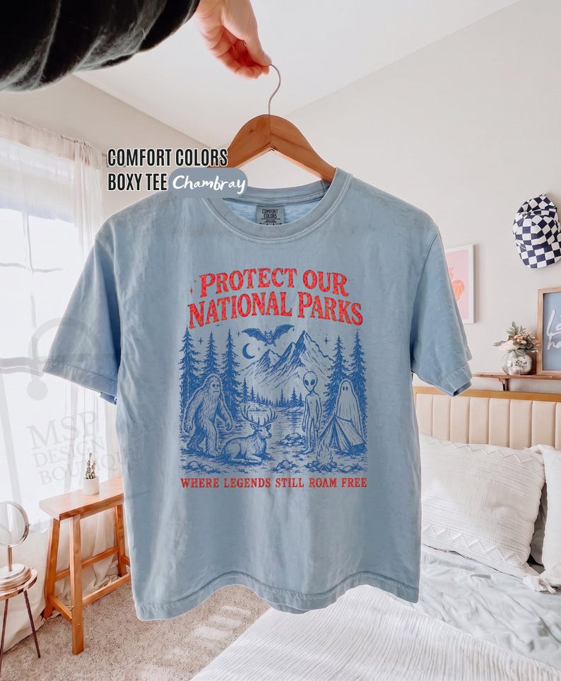 May include: A light blue short-sleeve t-shirt with a graphic print in red and blue. The text reads "PROTECT OUR NATIONAL PARKS" and "WHERE LEGENDS STILL ROAM FREE" above and below an illustration of mountains, a forest, a deer, a ghost, and an alien.