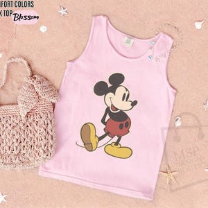 May include: A light pink tank top with a graphic of Mickey Mouse in black, red, and yellow. The words "COMFORT COLORS TANK TOP Blossom" are in the upper left corner. A pink woven handbag is on the left.