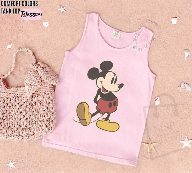 May include: A light pink tank top with a graphic of Mickey Mouse in black, red, and yellow. The words "COMFORT COLORS TANK TOP Blossom" are in the upper left corner. A pink woven handbag is on the left.