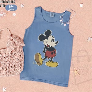 May include: A blue tank top featuring a classic Mickey Mouse graphic. The sleeveless shirt has a round neckline and is made of a soft material. The words "COMFORT COLORS" and "TANK TOP" are printed on the top left of the image, and the word "Blue Jean" is printed on a blue oval.