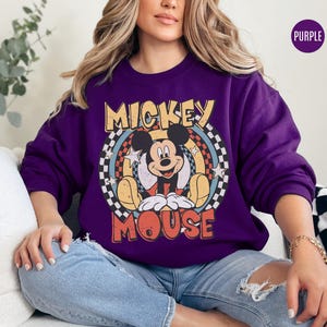 Puede incluir: Una sudadera morada con un gr&aacute;fico retro de Mickey Mouse. El dise&ntilde;o incluye las palabras "Mickey Mouse" en una fuente vintage, con un fondo circular de estrellas y rayas. La sudadera es de color morado liso.