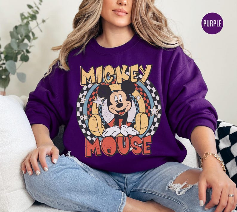 Puede incluir: Una sudadera morada con un gr&aacute;fico retro de Mickey Mouse. El dise&ntilde;o incluye las palabras "Mickey Mouse" en una fuente vintage, con un fondo circular de estrellas y rayas. La sudadera es de color morado liso.