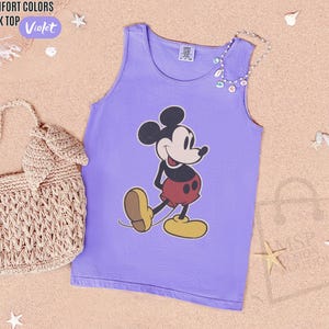 May include: Violet tank top featuring a classic Mickey Mouse graphic. The tank top is a light purple color and has a round neckline. The words "COMFORT COLORS TANK TOP" and the color "Violet" are printed on the top left corner.