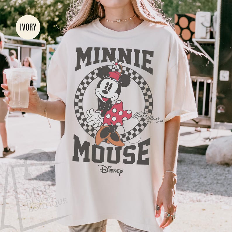 May include: Ivory-colored t-shirt featuring a graphic of Minnie Mouse. The design includes the text "MINNIE MOUSE" in a bold, dark gray font, with Minnie Mouse in the center. The shirt has a relaxed fit and short sleeves.