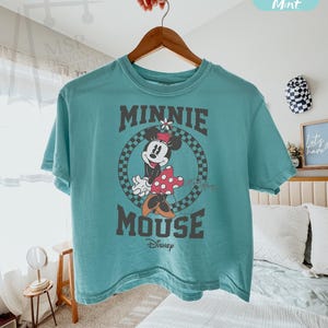 以下が含まれることがあります： ミニーマウスのグラフィックがプリントされた、ティール色のボクシーTシャツ。グラフィックは、市松模様の背景にミニーが描かれた円、"MINNIE MOUSE"の文字、ディズニーのロゴが特徴です。シャツは木製のハンガーに掛けられています。