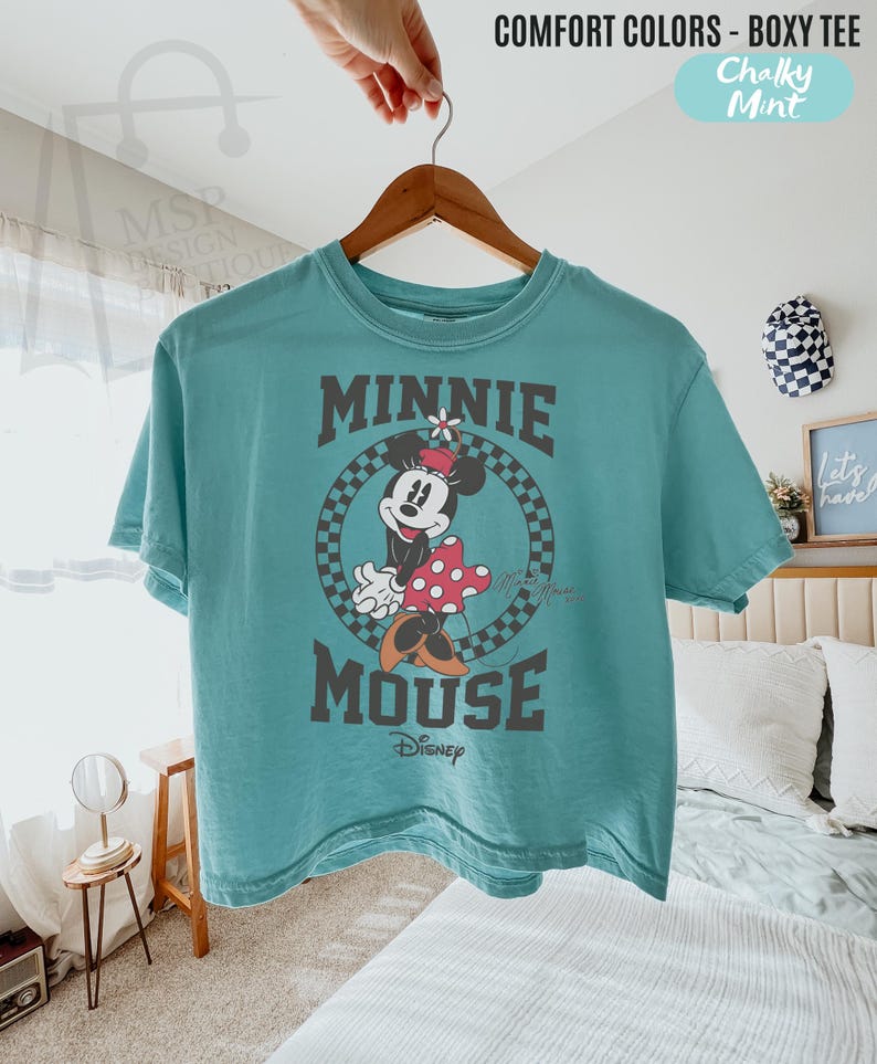 以下が含まれることがあります： ミニーマウスのグラフィックがプリントされた、ティール色のボクシーTシャツ。グラフィックは、市松模様の背景にミニーが描かれた円、"MINNIE MOUSE"の文字、ディズニーのロゴが特徴です。シャツは木製のハンガーに掛けられています。