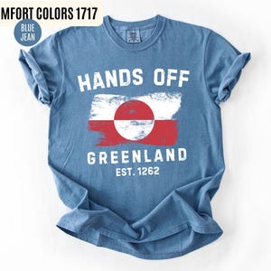 May include: Blue jean colored t-shirt with the words "HANDS OFF GREENLAND" in white block letters. The shirt features a Greenland flag design and the text "EST. 1262". The shirt is a Comfort Colors 1717.