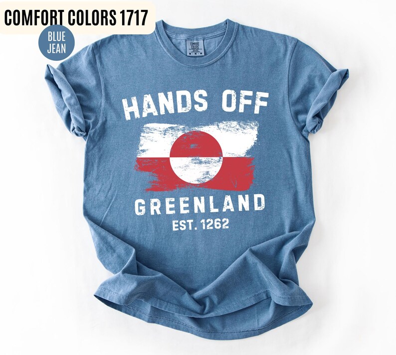 May include: Blue jean colored t-shirt with the words "HANDS OFF GREENLAND" in white block letters. The shirt features a Greenland flag design and the text "EST. 1262". The shirt is a Comfort Colors 1717.
