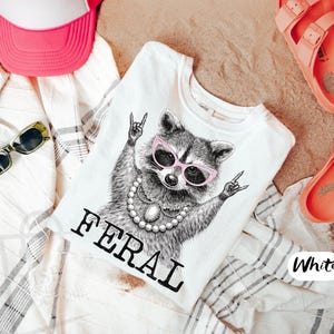 May include: White t-shirt with a graphic of a raccoon wearing pink sunglasses and a pearl necklace, with the word "FERAL" in black. The shirt is surrounded by a pink hat, sunglasses, and coral sandals.