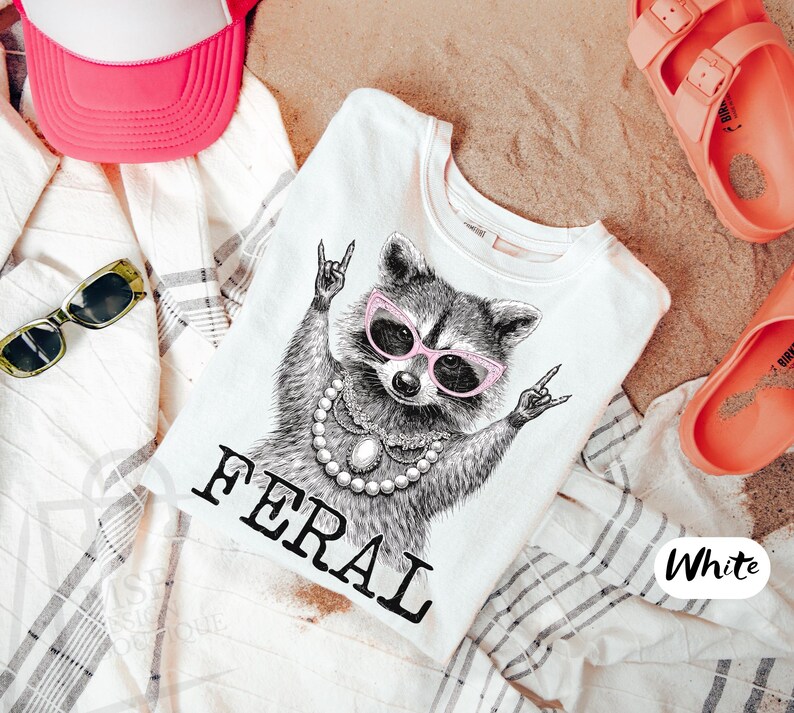 May include: White t-shirt with a graphic of a raccoon wearing pink sunglasses and a pearl necklace, with the word "FERAL" in black. The shirt is surrounded by a pink hat, sunglasses, and coral sandals.