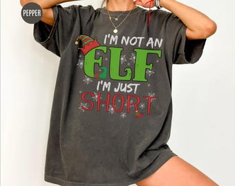 May include: Dark gray t-shirt with the text "I'M NOT AN ELF I'M JUST SHORT" in green and red. The design includes an elf hat and snowflakes. The shirt is a relaxed fit.