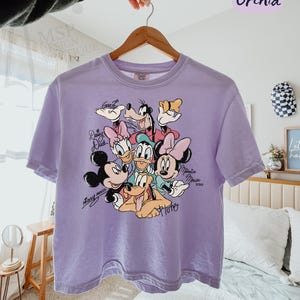 Puede incluir: Una camiseta holgada de color lila claro con un gr&aacute;fico de Mickey Mouse, Minnie Mouse, Donald Duck, Daisy Duck, Goofy y Pluto. La camiseta tiene las palabras "COMFORT COLORS - BOXY TEE" y "Orchid" impresas en la parte superior.
