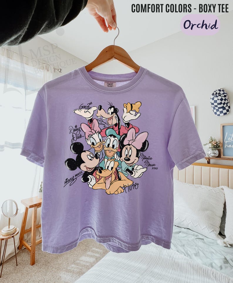 Puede incluir: Una camiseta holgada de color lila claro con un gr&aacute;fico de Mickey Mouse, Minnie Mouse, Donald Duck, Daisy Duck, Goofy y Pluto. La camiseta tiene las palabras "COMFORT COLORS - BOXY TEE" y "Orchid" impresas en la parte superior.
