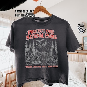 May include: A charcoal gray boxy t-shirt featuring a graphic print with the text "PROTECT OUR NATIONAL PARKS" and "WHERE LEGENDS STILL ROAM FREE" in red. The illustration depicts a forest, mountains, a bat, a deer, a ghost, and an alien.